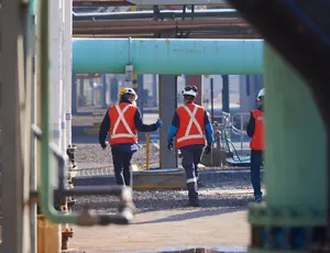 대형 파이프라인이 설치된 산업 현장에서 안전 조끼와 보호 장비를 착용한 작업자 3명이 시설을 점검하며 이동하는 모습. 석유화학 또는 수처리 플랜트로 보이며, 산업 안전 및 인프라가 강조된 장면.